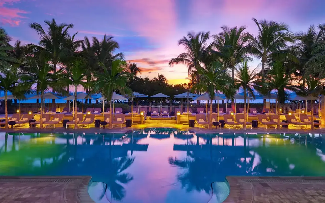 A serene pool scene at sunset with lounge chairs, umbrellas, and palm trees.