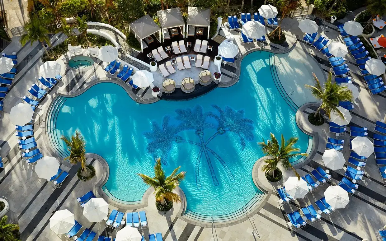 An aerial view of a pool area with a blue pool, surrounded by blue lounge chairs and white umbrellas, with palm trees and a concrete deck.