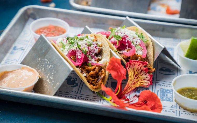 Two crispy meat tacos with fresh pickled onions, queso fresco, fresh garnishes and side toppings for tacos