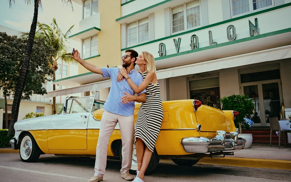 Couple taking selfies by a classic car in front of the Avalon Hotel on Ocean Drive
