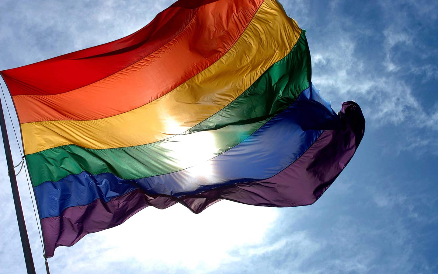 LGBTQ+ Flag flying in blue skies with bright sun shining through